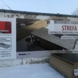 Billboard reklamowy z wizualizacją nowoczesnej kuchni i informacją o materiałach do produkcji mebli, umieszczony na zniszczonej ścianie budynku, obok stosu płyt drewnianych w zimowym otoczeniu.