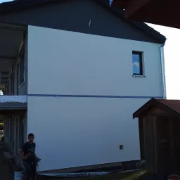 Elewacja domu jednorodzinnego w trakcie malowania natryskowego, widoczny pracownik z pistoletem malarskim, część elewacji w kolorze szarym, druga część w stanie surowym, małe okno z odbiciem nieba.