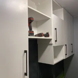 Białe szafki wiszące z czarnymi uchwytami, na półce w szafce wiertarka akumulatorowa i wkrętarka, czarna ściana pod szafkami, widoczna instalacja.