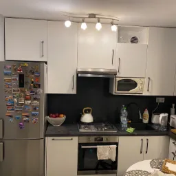 Wnętrze kuchni z białymi szafkami, srebrną lodówką ozdobioną magnesami, czarnym panelem nad blatem i okrągłym stołem z dekoracjami.