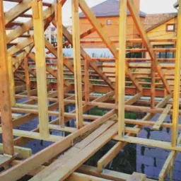Konstrukcja drewniana dachu w trakcie budowy, widoczne elementy więźby i ściany domu w tle.