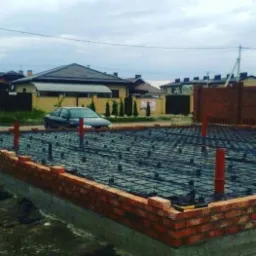 Fundament domu jednorodzinnego z obwodem z cegły, widoczna siatka zbrojeniowa i pionowe rury instalacyjne, w tle zabudowa jednorodzinna i samochód.
