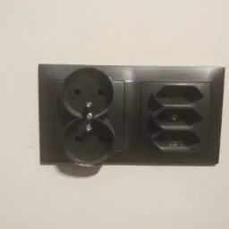 Podwójne, ciemne gniazdko elektryczne z uziemieniem typu francuskiego i niemieckiego, zamontowane na jasnej ścianie. Widoczne ślady montażu.