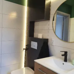 Nowoczesna łazienka z białymi płytkami, czarnym akcentem w postaci zabudowy WC i umywalki, okrągłym lustrem w złotej ramie, podświetleniem LED i zielonym sufitem.