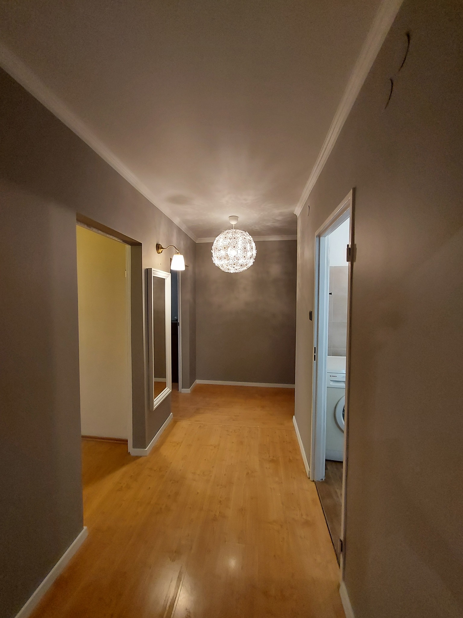 Korytarz w mieszkaniu po odświeżeniu: szare ściany, drewniana podłoga, ozdobna lampa sufitowa, lustro z kinkietem i widok na łazienkę z pralką.