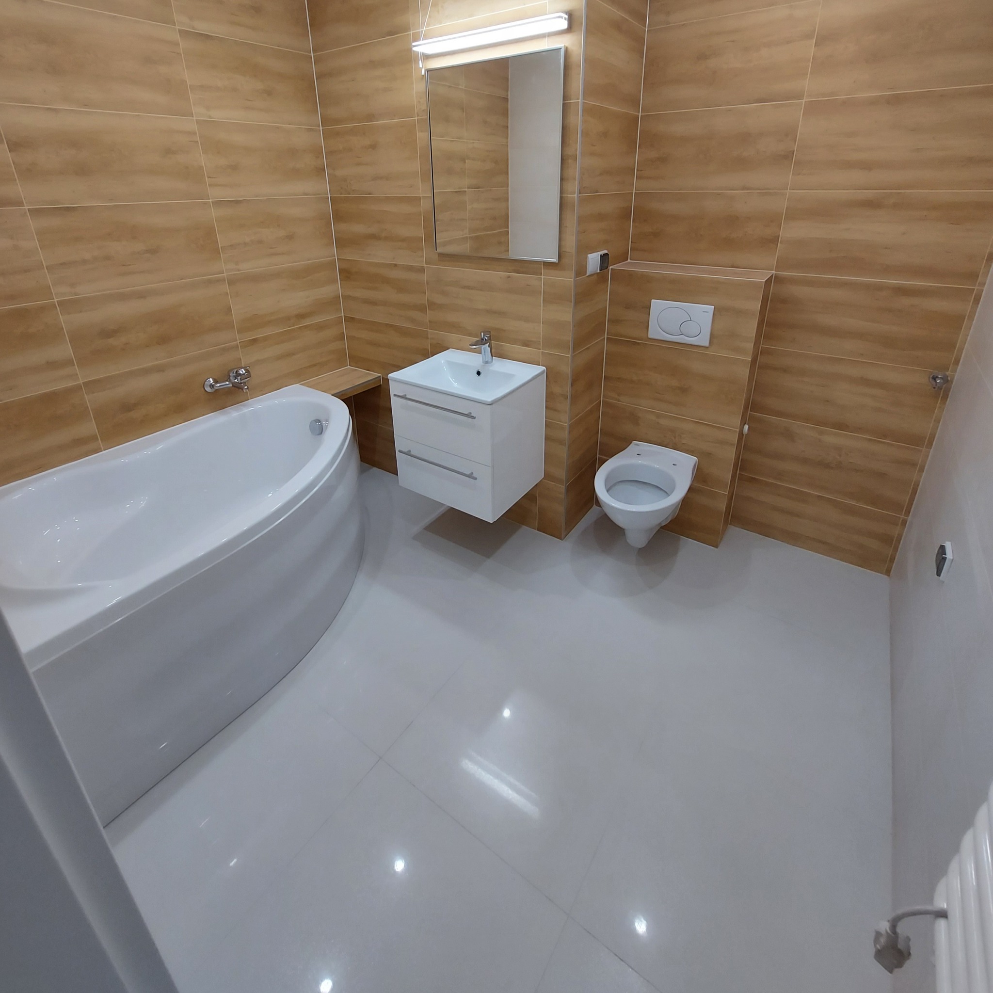 Nowoczesna łazienka z beżowymi płytkami ściennymi imitującymi drewno, białą wanną o zaokrąglonym kształcie, podwieszaną szafką z umywalką i wiszącą toaletą; oświetlenie punktowe nad lustrem.