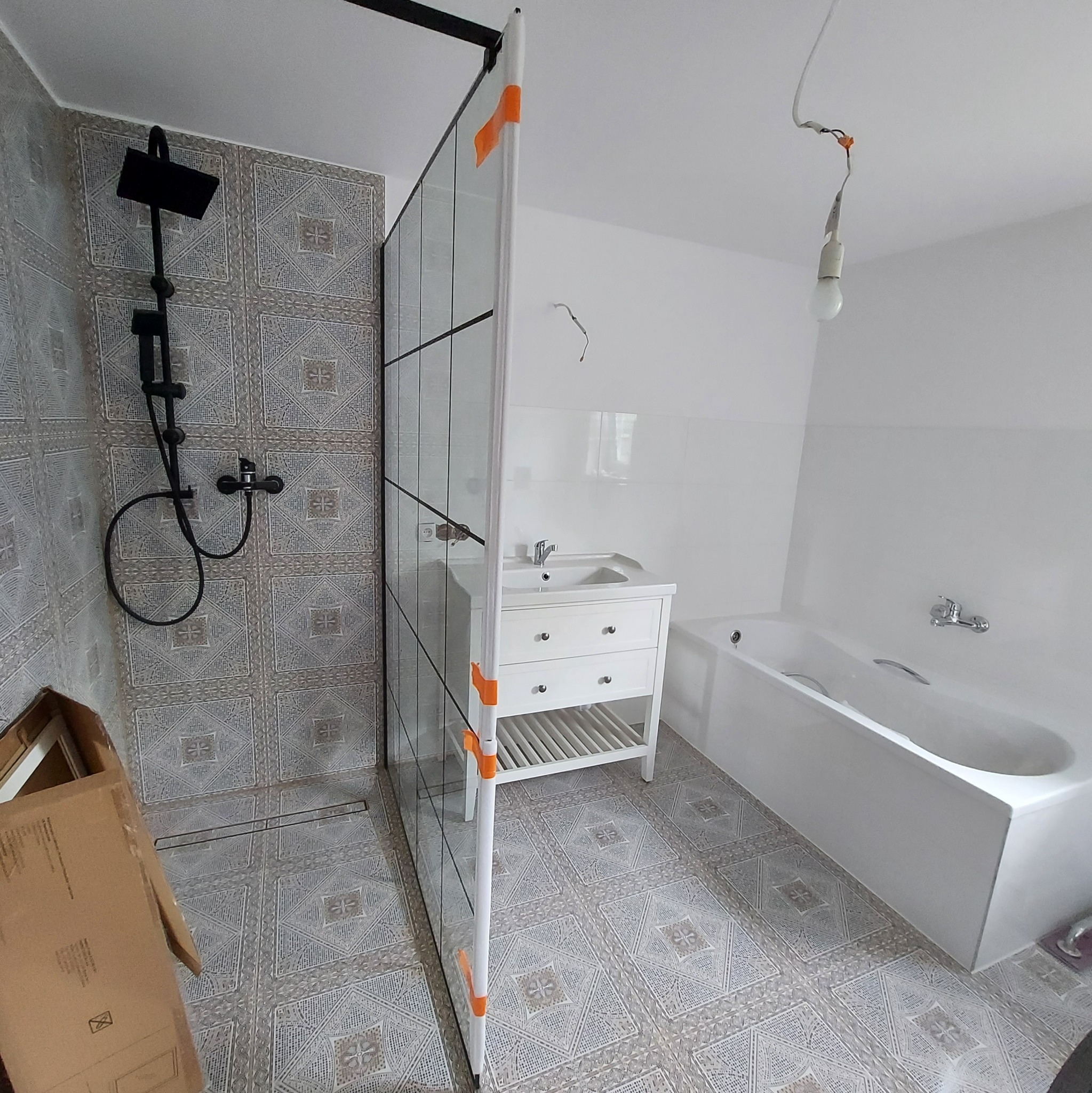 Wnętrze łazienki w trakcie prac: czarny zestaw prysznicowy na ścianie z wzorzystymi płytkami, szklana kabina prysznicowa z pomarańczowymi zabezpieczeniami, biała umywalka z szafką, wanna i wisząca...