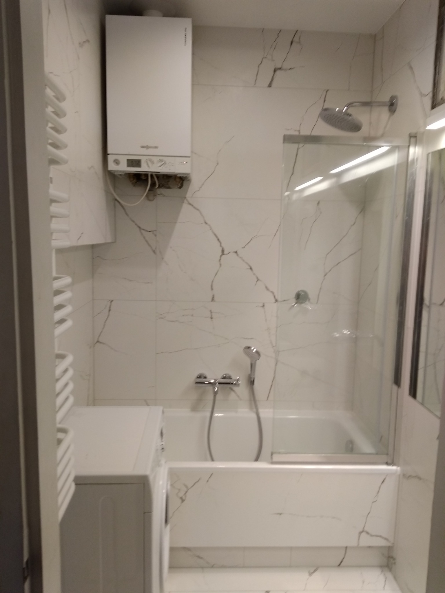 Biała łazienka z wanną za szklaną szybą, pralką obok grzejnika drabinkowego i piecem gazowym na ścianie wyłożonej płytkami imitującymi marmur.