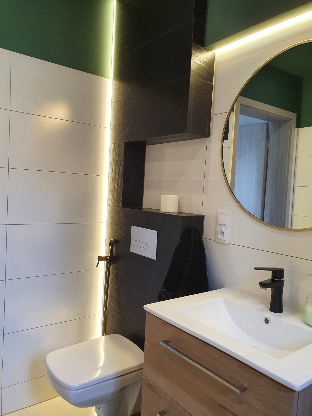 Nowoczesna łazienka z białymi płytkami, czarnym akcentem w postaci zabudowy WC i umywalki, okrągłym lustrem w złotej ramie, podświetleniem LED i zielonym sufitem.