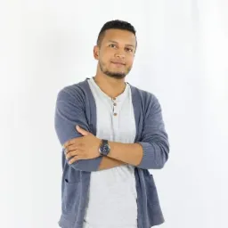 Mężczyzna o ciemnej karnacji, ubrany w szary t-shirt i granatowy kardigan, stoi na białym tle z założonymi rękami, prezentując zegarek na skórzanym pasku.