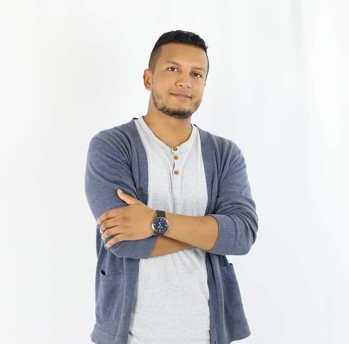 Mężczyzna o ciemnej karnacji, ubrany w szary t-shirt i granatowy kardigan, stoi na białym tle z założonymi rękami, prezentując zegarek na skórzanym pasku.