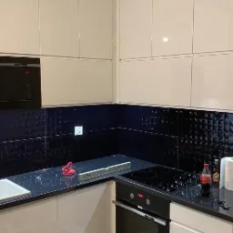 Częściowo wykończona kuchnia z błyszczącymi białymi szafkami, czarnym blatem z poziomnicą, czarną mozaiką na ścianie i wbudowaną płytą grzewczą.