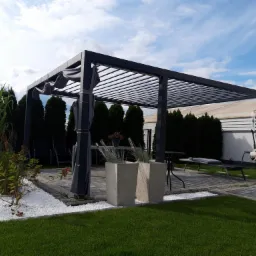 Nowoczesna pergola z metalową konstrukcją, zasłonami, betonowymi donicami i białymi kamykami na trawniku.