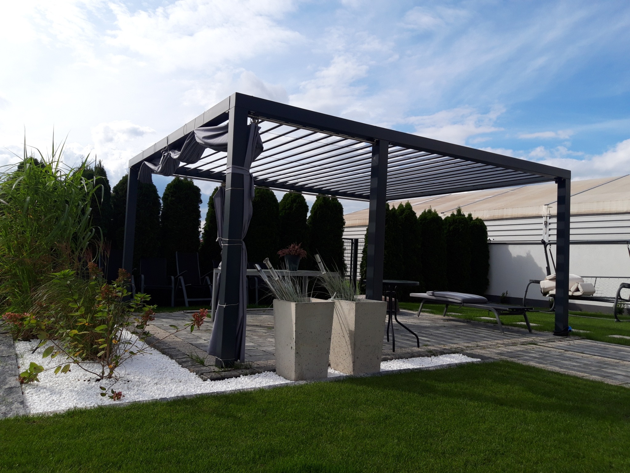 Nowoczesna pergola z metalową konstrukcją, zasłonami, betonowymi donicami i białymi kamykami na trawniku.
