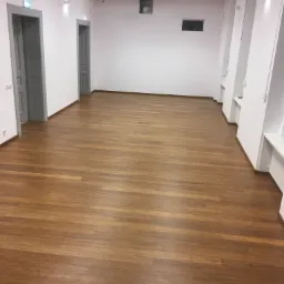 Wnętrze przestronnego pomieszczenia z nową, błyszczącą podłogą z desek w odcieniach brązu, białymi ścianami z szarymi drzwiami i drewnianymi belkami stropowymi.