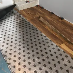 Proces układania paneli podłogowych: widoczna izolacja akustyczna z nadrukiem, częściowo ułożone panele z brązowym wzorem drewna, kliny dystansowe przy ścianie i paczka paneli przy schodach.