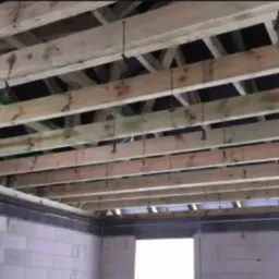 Sufit poddasza z drewnianymi belkami i elementami mocującymi, widok na surową konstrukcję z bloków betonowych i otwór okienny.