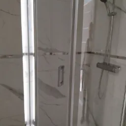 Kabina prysznicowa z białymi marmurkowymi płytkami, widoczne srebrne okucia, bateria termostatyczna i deszczownica, odbicie osoby w szybie.