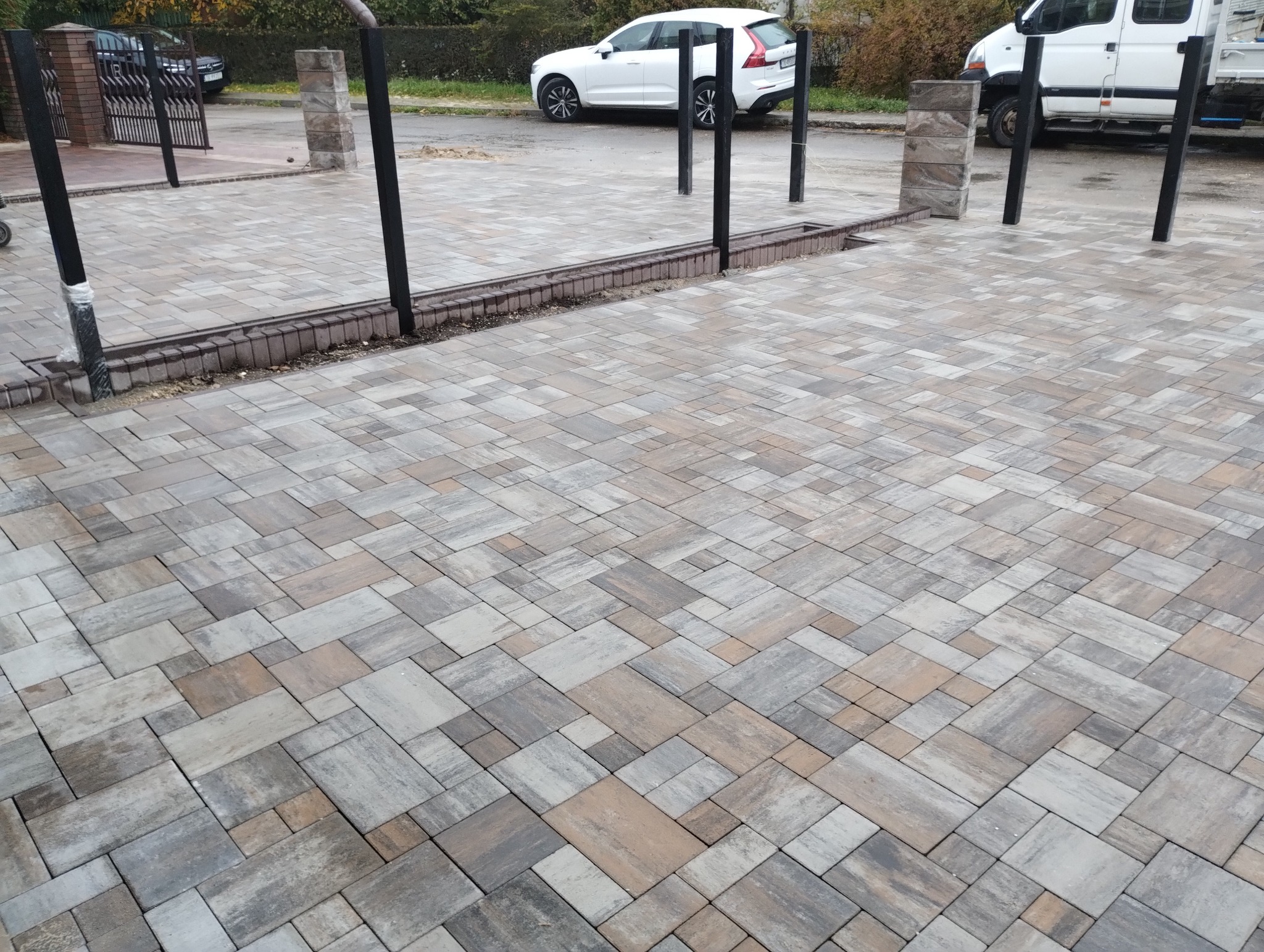 Ułożona kostka brukowa w odcieniach szarości i beżu, tworząca wzór na podjeździe. Widoczne słupki ogrodzeniowe i samochody w tle. Całość w neutralnej tonacji.
