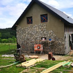 Artur Rosinski budownictwo - Budynek w trakcie wykańczania elewacji kamiennej, widoczne rusztowania, częściowo położony kamień elewacyjny, zabezpieczone okna folią, w tle zalesione wzgórza.