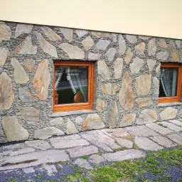 Elewacja domu wykończona nieregularnym kamieniem naturalnym, z widocznymi dwoma oknami z drewnianymi ramami i fragmentem kamiennego chodnika.