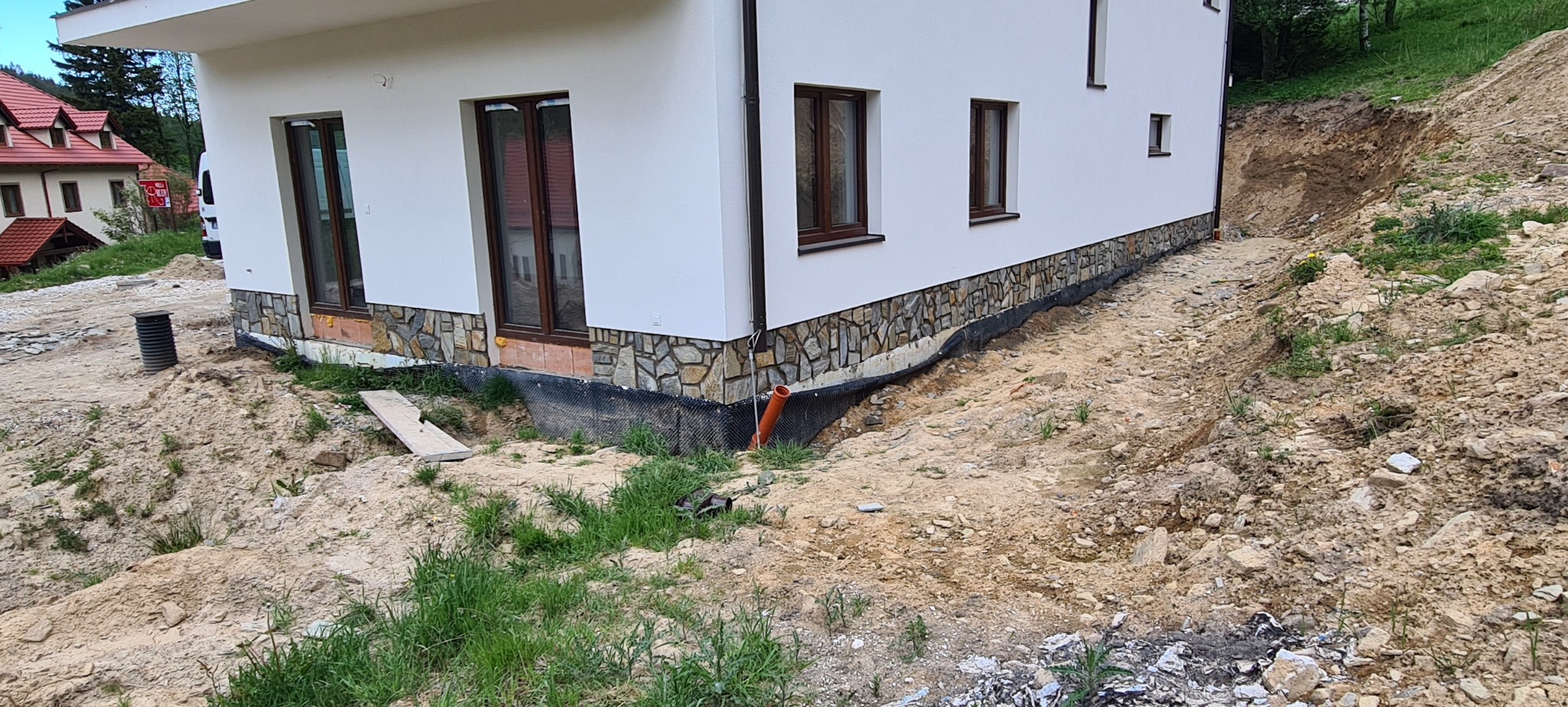 Wykończona elewacja domu z kamienną okładziną, widoczna hydroizolacja fundamentów i teren po pracach ziemnych.