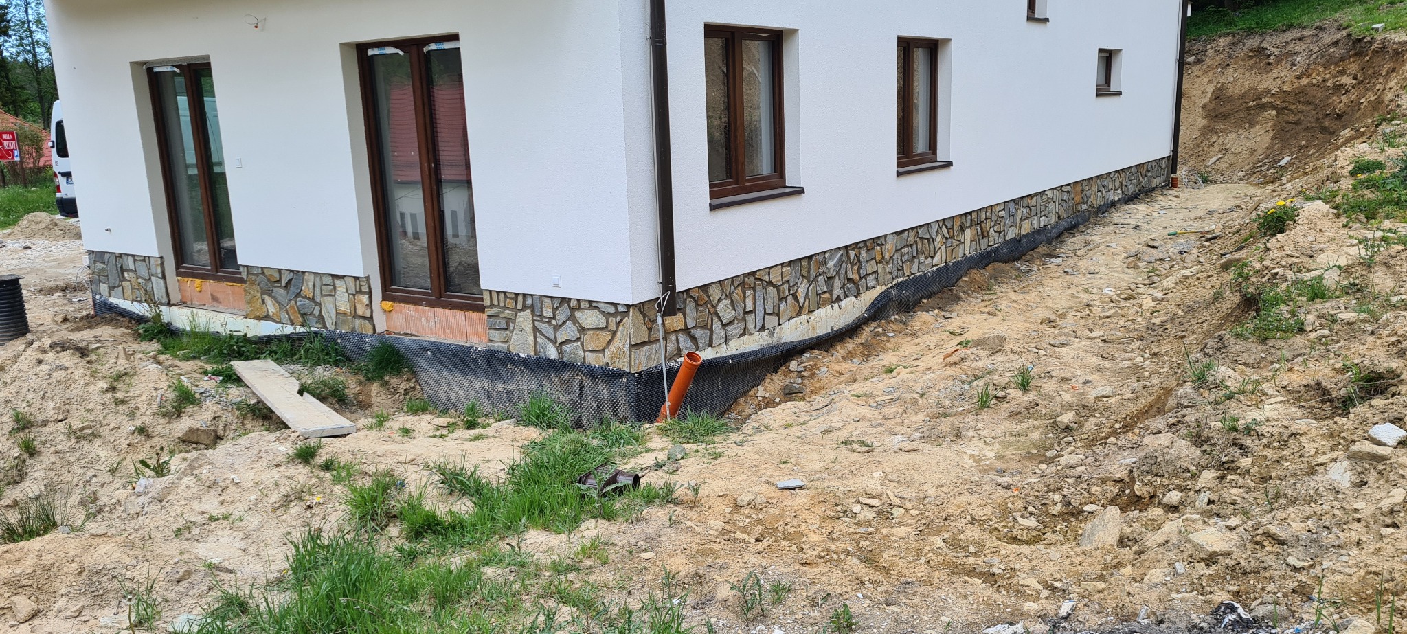 Wykończenie elewacji domu jednorodzinnego kamieniem dekoracyjnym na fundamencie, widoczne okna z brązowymi ramami i teren wokół w trakcie prac ziemnych.