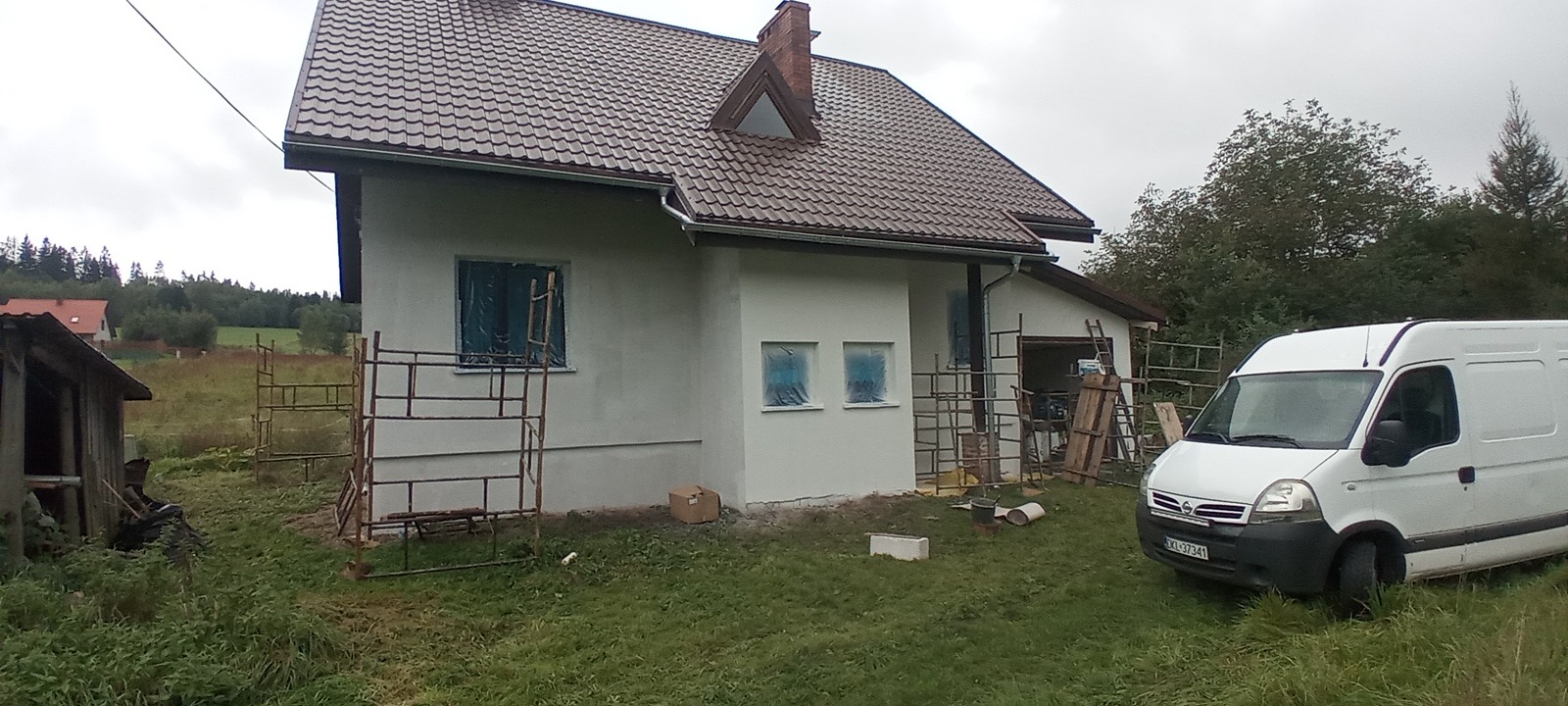 Dom jednorodzinny po nałożeniu jasnego tynku elewacyjnego, z zabezpieczonymi oknami folią, rusztowania rozstawione wokół, biały bus zaparkowany obok.