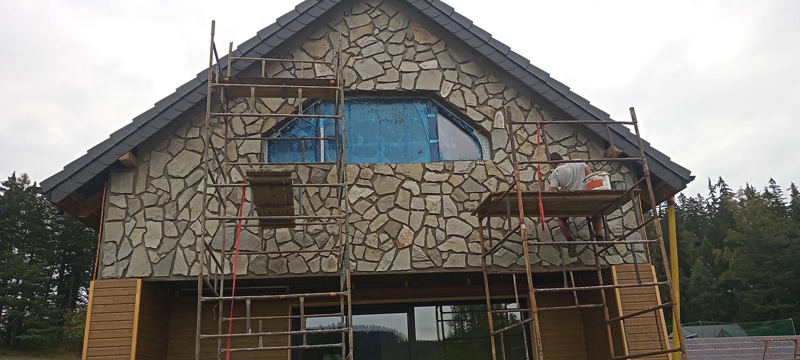 Dom z elewacją z kamienia w trakcie budowy, widoczne rusztowania i pracownik z wiadrem farby.