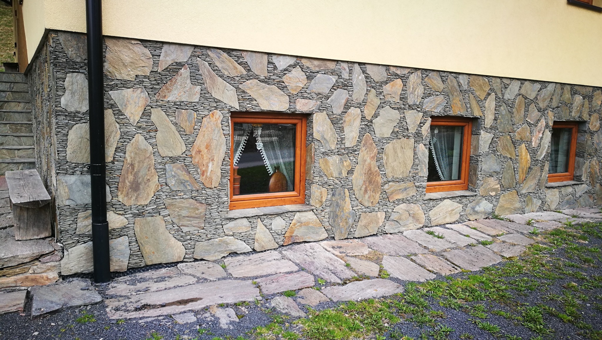 Elewacja domu wykończona nieregularnym kamieniem naturalnym, z widocznymi dwoma oknami z drewnianymi ramami i fragmentem kamiennego chodnika.