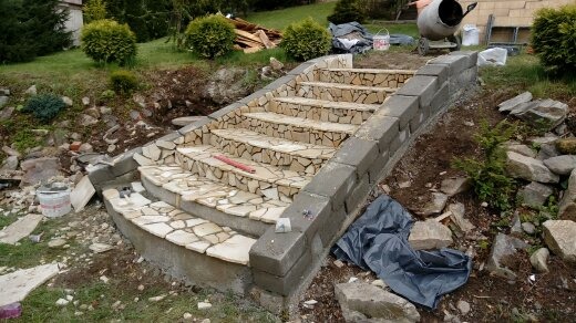 Kamienne schody zewnętrzne w trakcie budowy, z widocznymi blokami betonowymi po bokach i mozaiką z kamienia naturalnego na stopniach, na tle ogrodu.