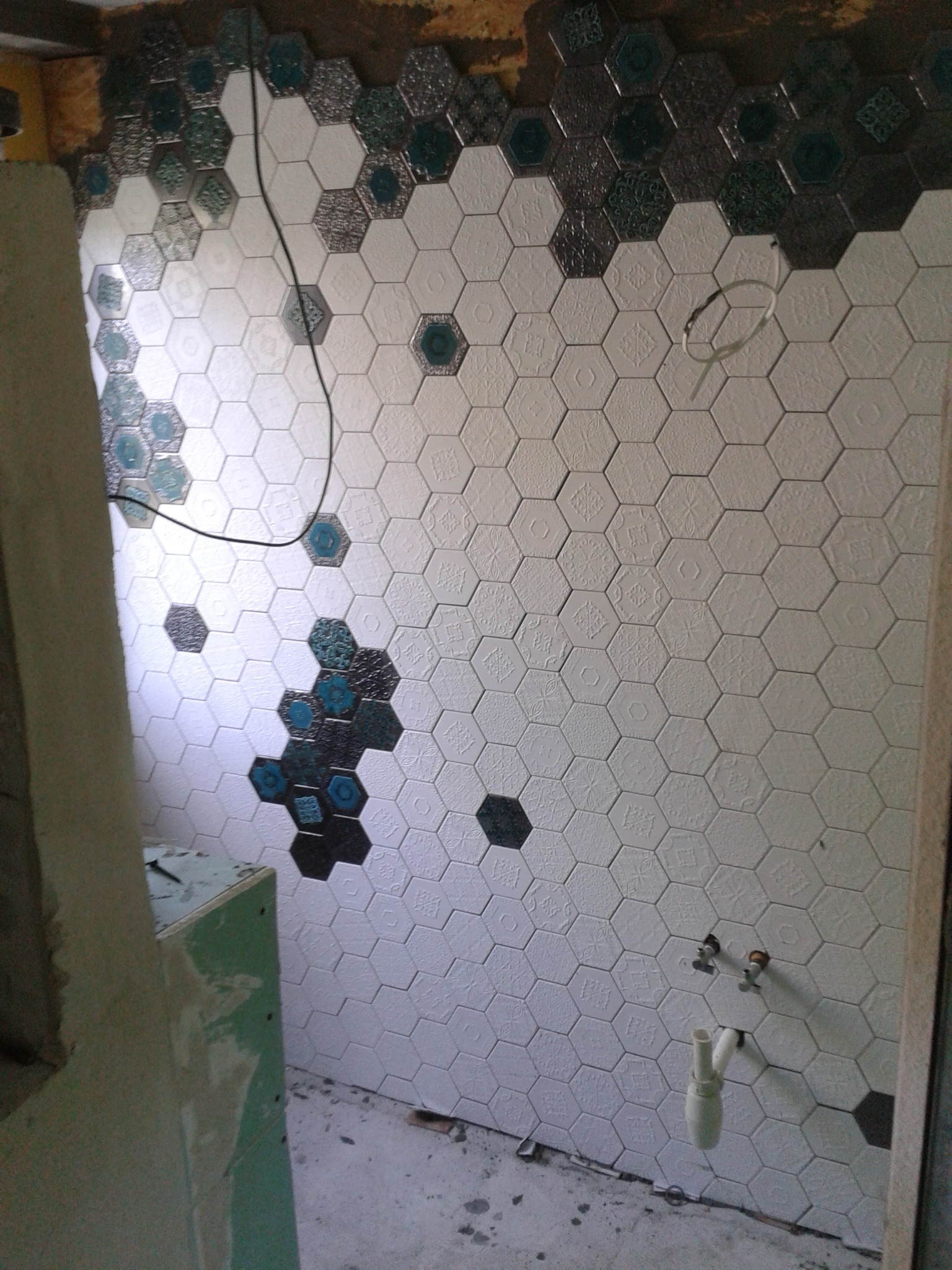 Ściana w łazience wyłożona heksagonalnymi płytkami z wytłaczanymi wzorami, w trakcie układania, z widocznymi przyłączami wodnymi i fragmentami nieukończonej górnej części oraz dolnej krawędzi.