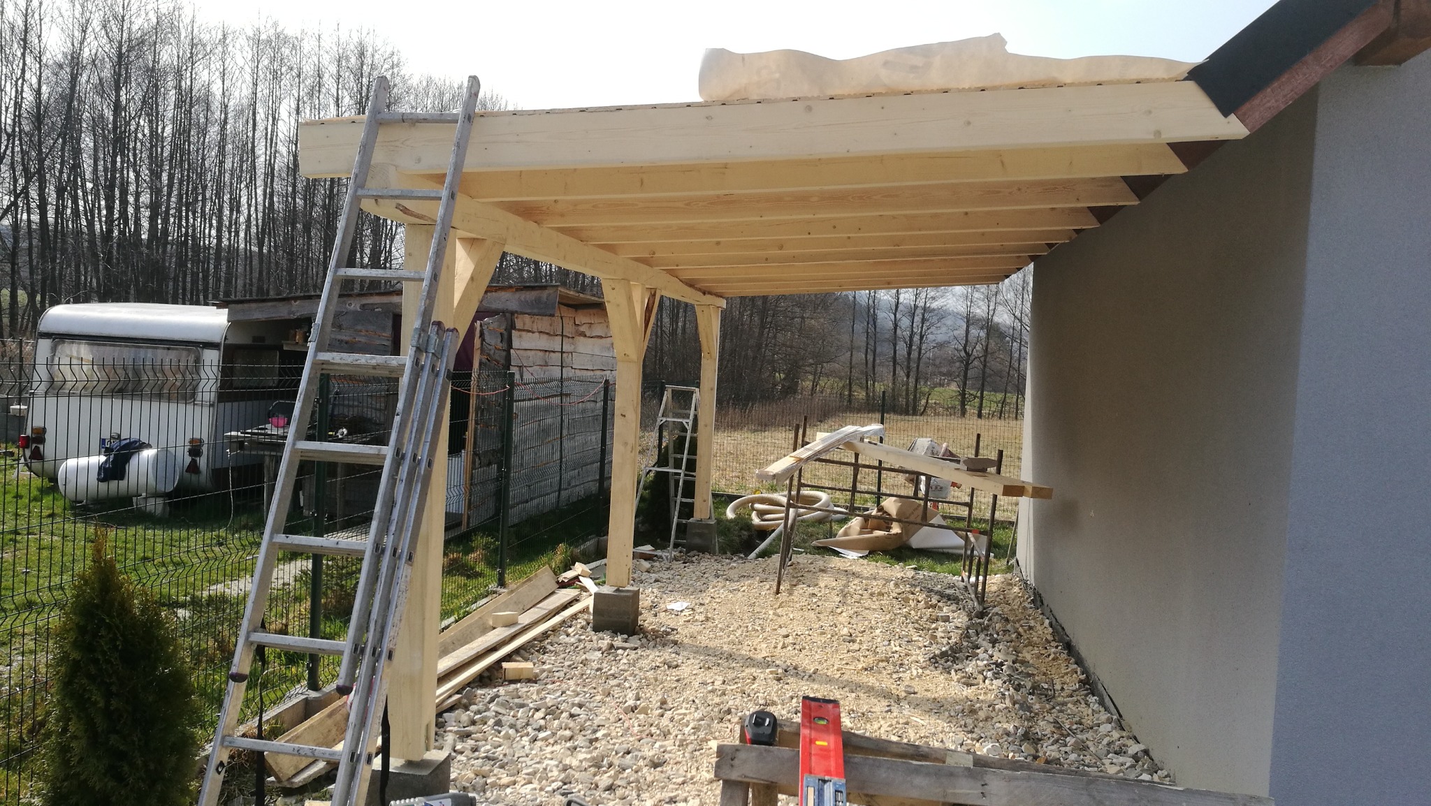 Wznoszenie drewnianej konstrukcji zadaszenia przylegającego do budynku, widoczne rusztowania, drabina oparta o konstrukcję, teren wokół wysypany żwirem, w tle przyczepa kempingowa i drzewa.