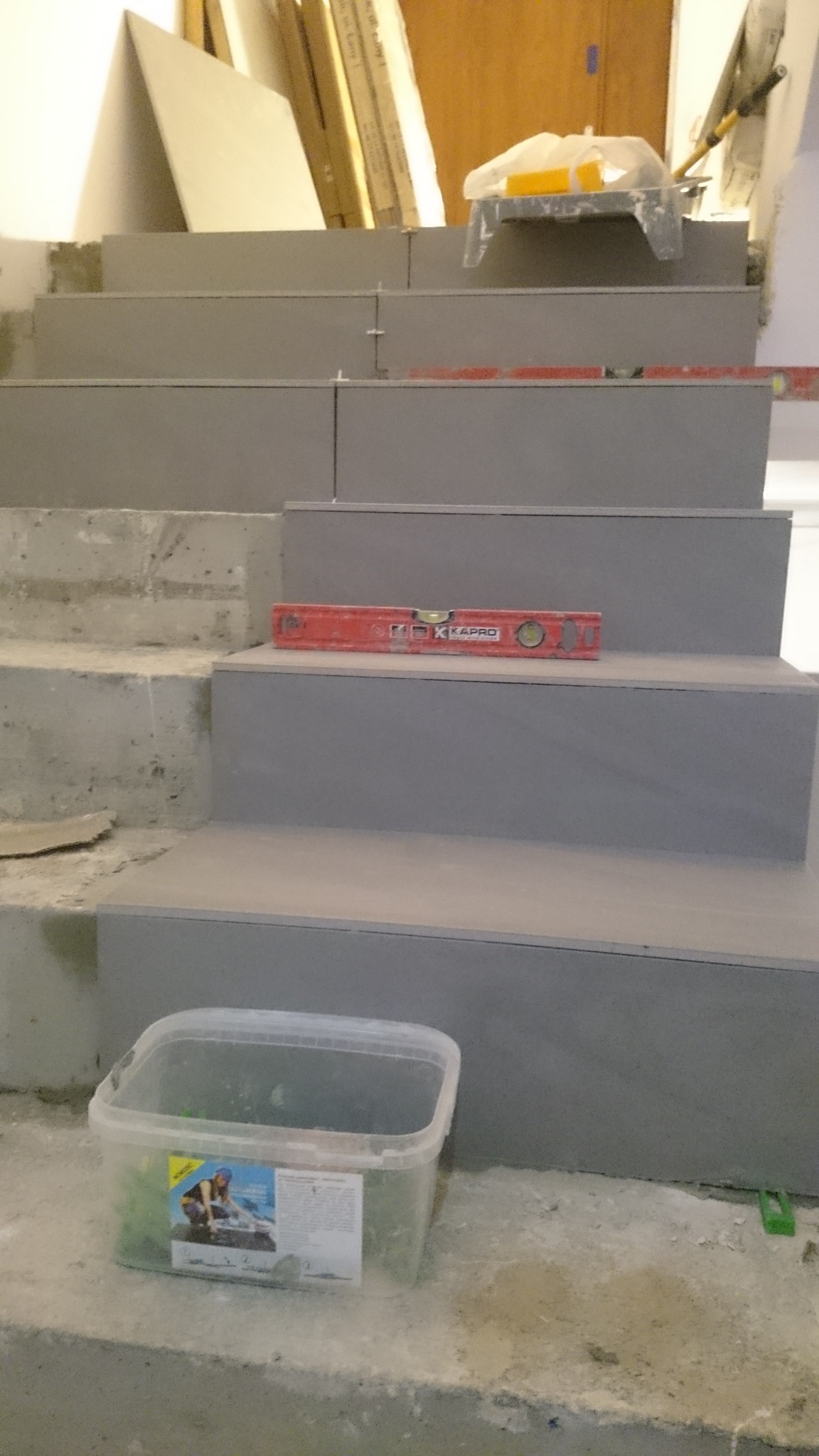 Schody w trakcie wykańczania płytkami, widoczna poziomica Kapro, plastikowe krzyżyki dystansowe i wiadro z zaprawą.