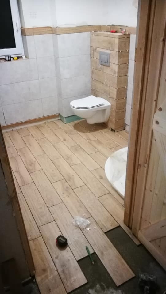 Łazienka w trakcie układania płytek podłogowych imitujących drewno, z częściowo obudowaną toaletą podwieszaną płytkami w podobnym odcieniu. Widoczne narzędzia: miarka i ołówek.
