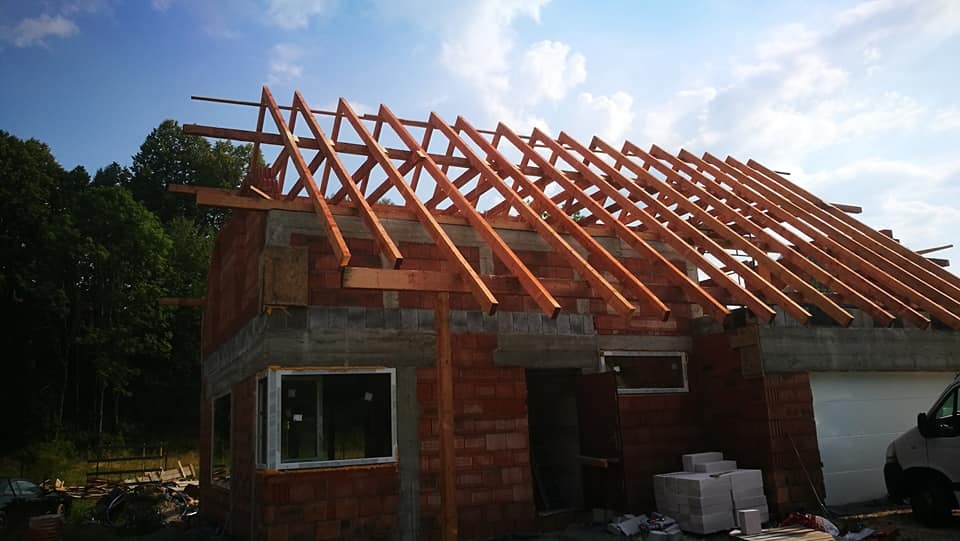 Budowa domu jednorodzinnego w stanie surowym otwartym: ściany z czerwonej cegły, wstawione okno z białymi ramami oraz konstrukcja drewniana dachu na tle błękitnego nieba z chmurami.