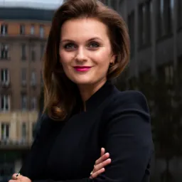 Portret uśmiechniętej kobiety w czarnym żakiecie, z założonymi rękami, na tle miejskiej zabudowy z budynkami o zróżnicowanej architekturze.