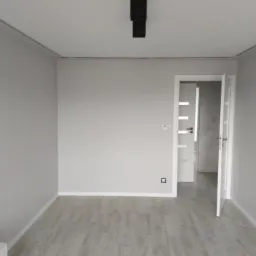 Pomalowane na szaro wnętrze pokoju z panelami podłogowymi, widoczna otwarta biała futryna drzwiowa i czarna lampa sufitowa.