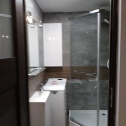 Marko wykonczenia wnętrz - Widok na łazienkę z kabiną prysznicową za szklanymi drzwiami, umywalką z szafką, toaletą i grzejnikiem drabinkowym, całość w szaro-białej kolorystyce z oświetleniem sufitowym.