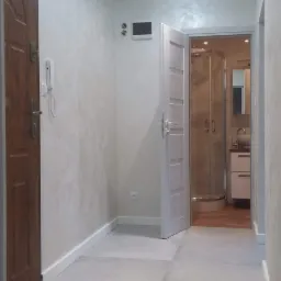 Korytarz po remoncie z szarą podłogą z płytek, białymi ścianami z efektem dekoracyjnym i otwartymi drzwiami do łazienki z kabiną prysznicową.