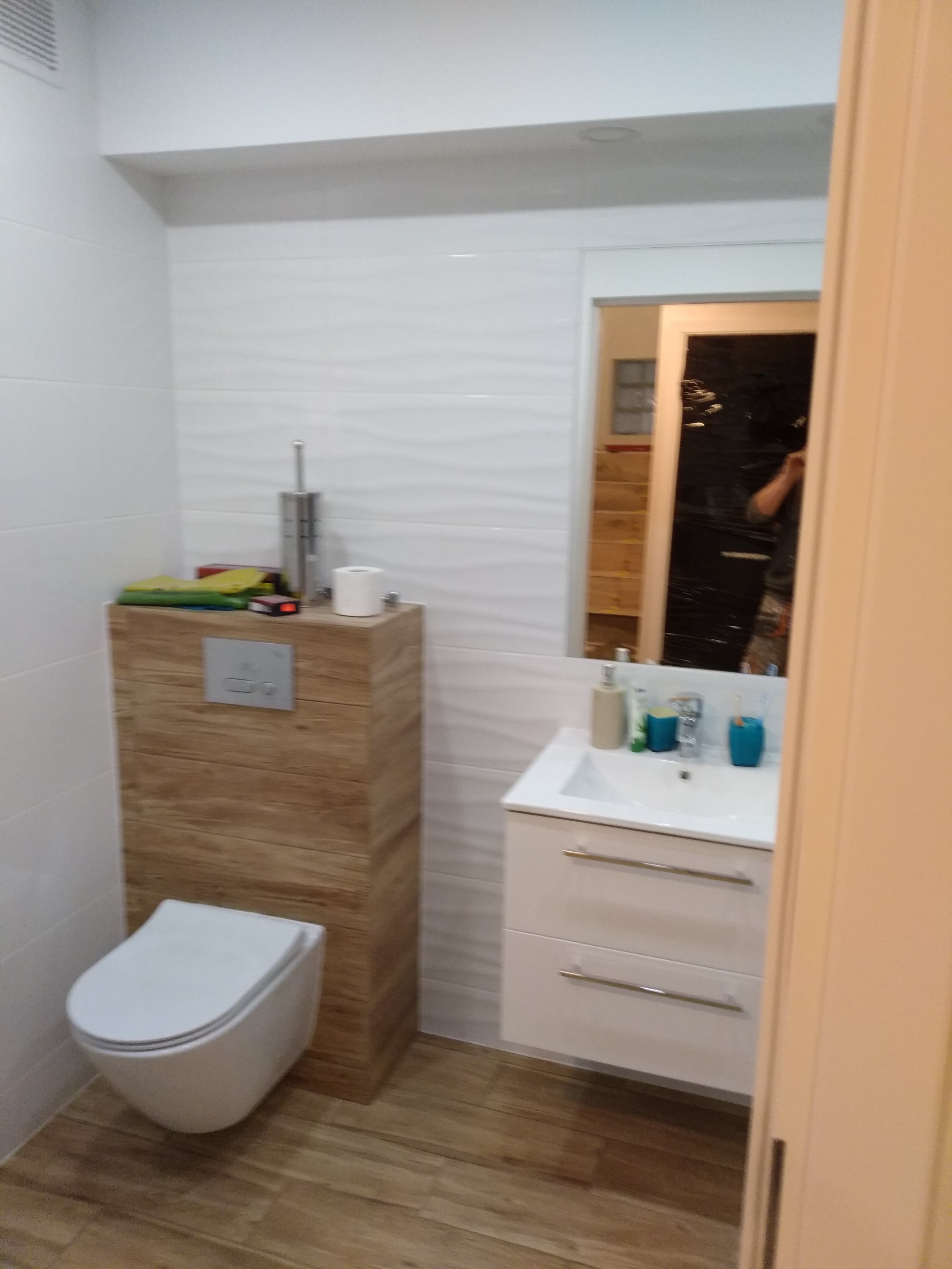 Nowoczesna łazienka z podwieszaną toaletą w obudowie z drewnopodobnych paneli, białą umywalką z szafką i lustrem, wykończona białymi płytkami z delikatnym wzorem fal.