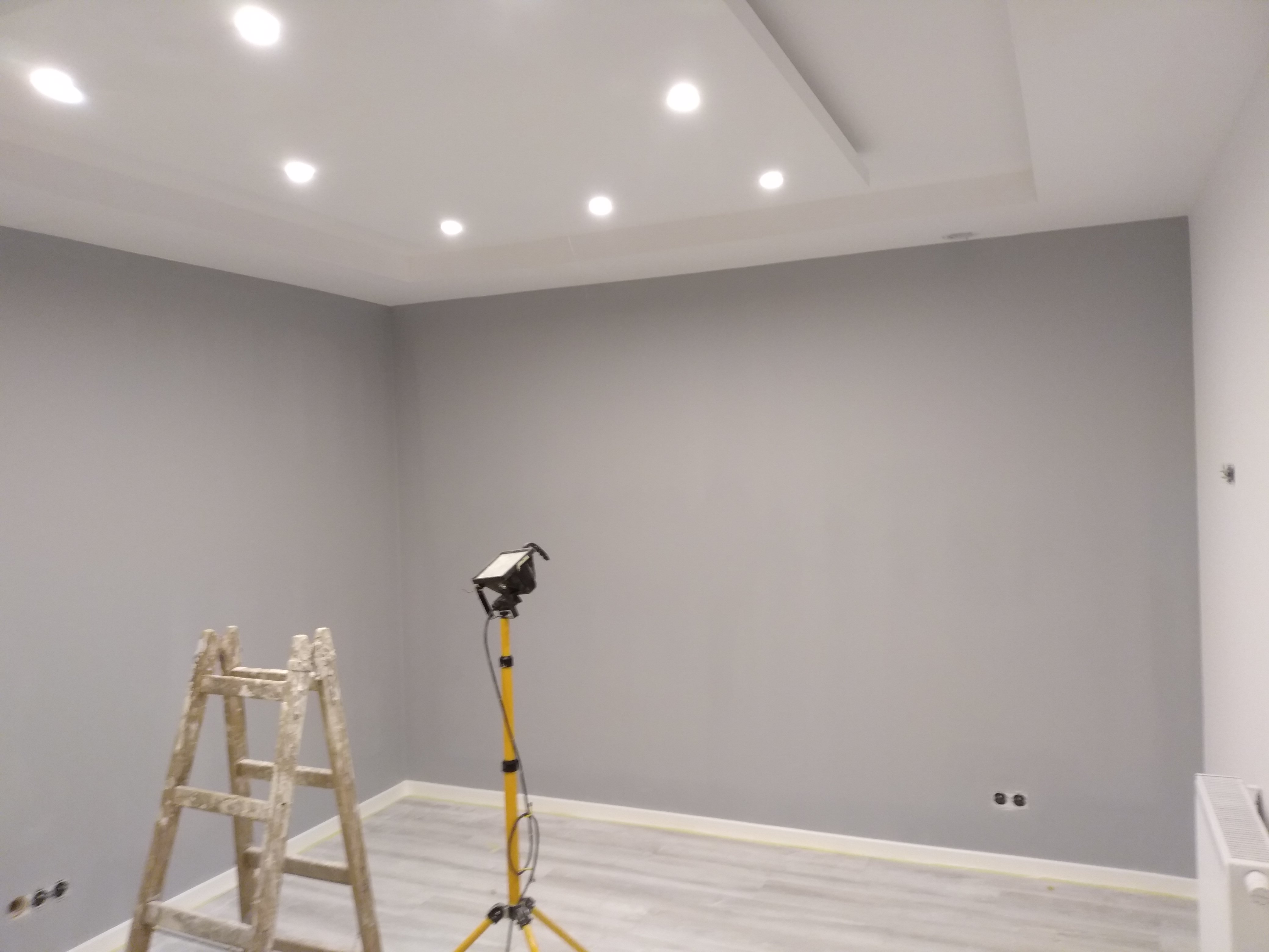 Świeżo pomalowane na szaro wnętrze pokoju z drewnianą drabiną i lampą budowlaną na trójnogu, widoczne gniazdka elektryczne i grzejnik.
