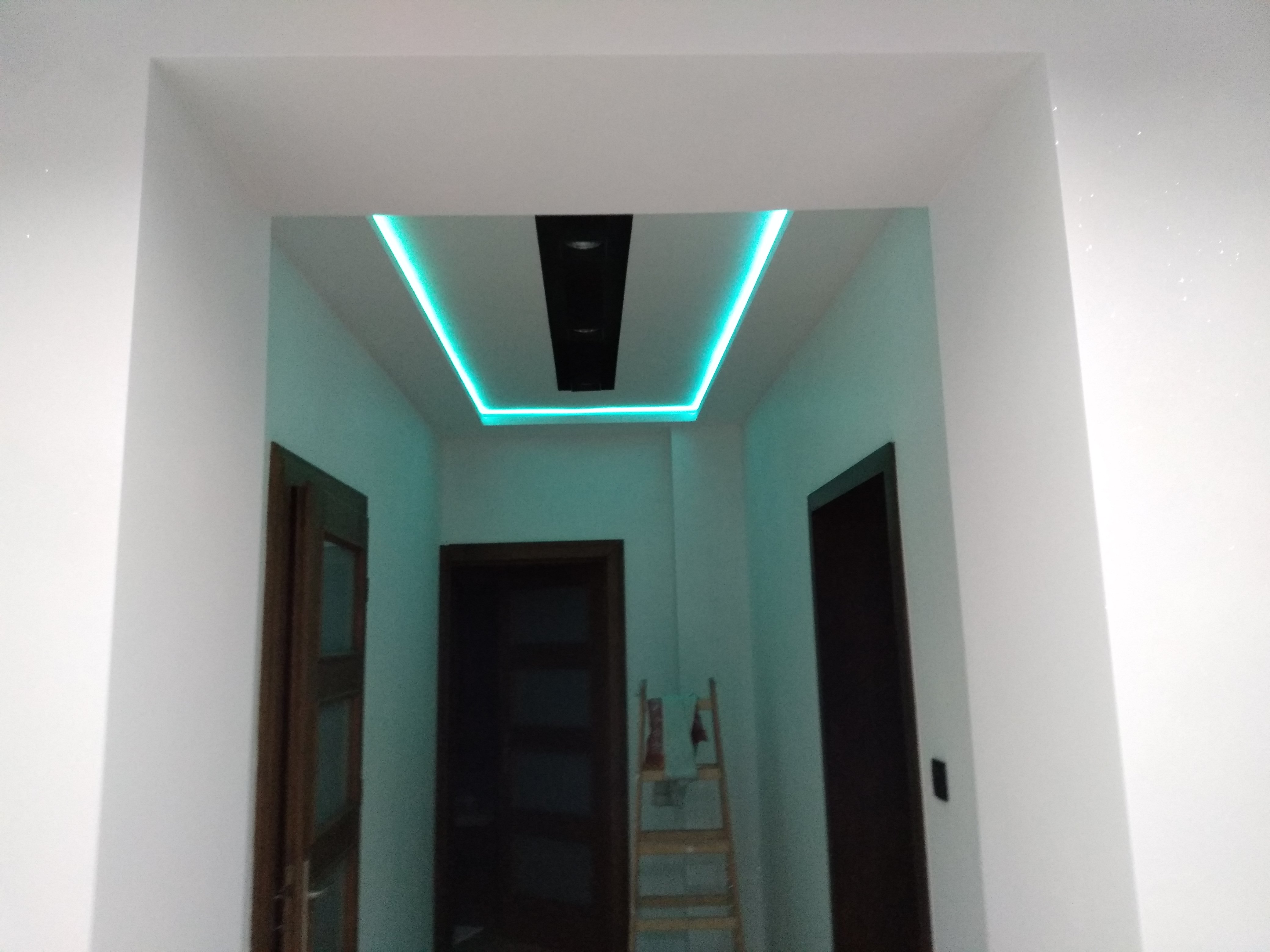Nowoczesny korytarz z podświetlanym na turkusowo sufitem napinanym i czarnym panelem centralnym z oświetleniem punktowym, widok na dwie ciemne drewniane ościeżnice drzwiowe i drabinę opartą o ścianę.
