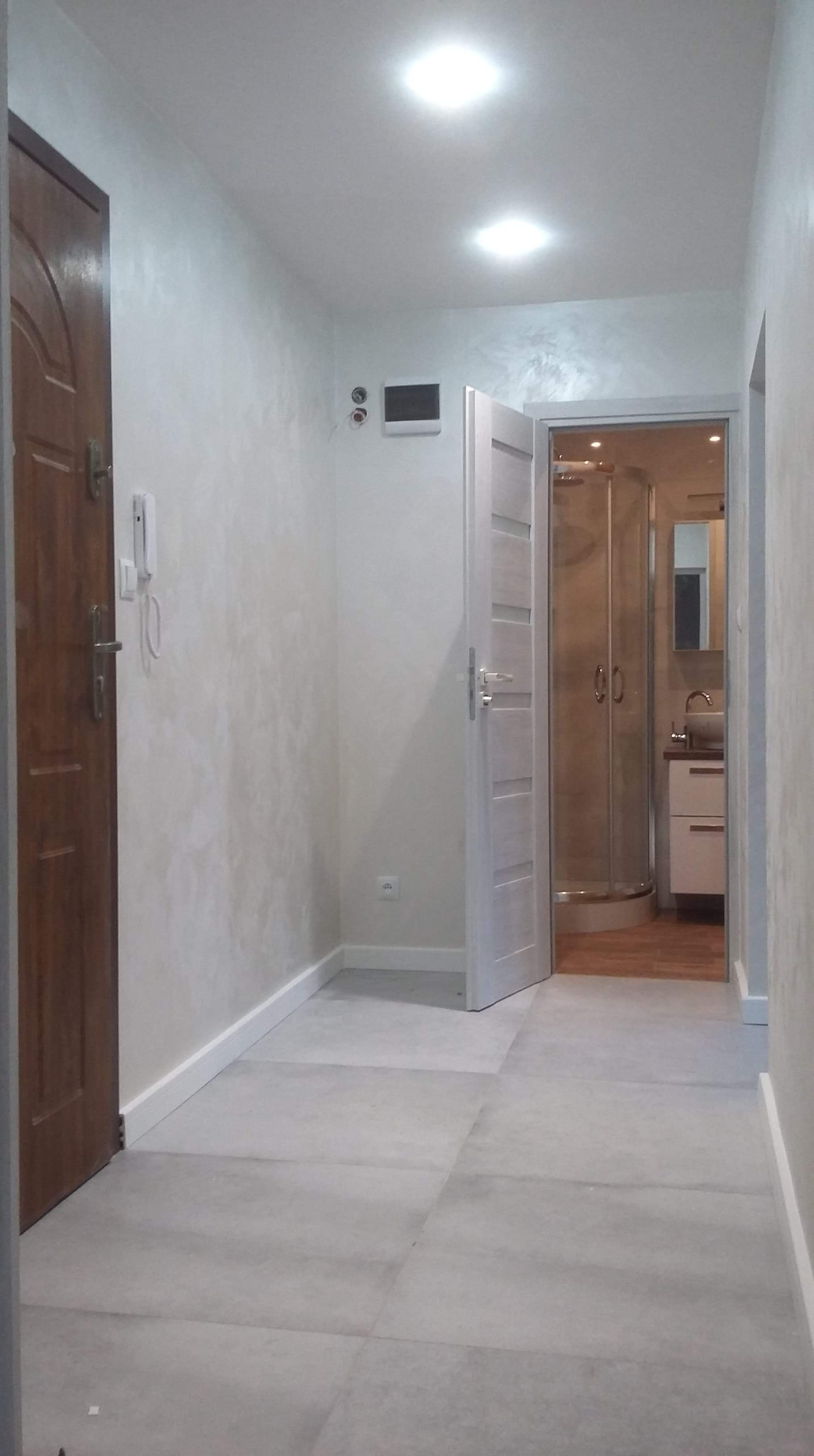 Korytarz po remoncie z szarą podłogą z płytek, białymi ścianami z efektem dekoracyjnym i otwartymi drzwiami do łazienki z kabiną prysznicową.