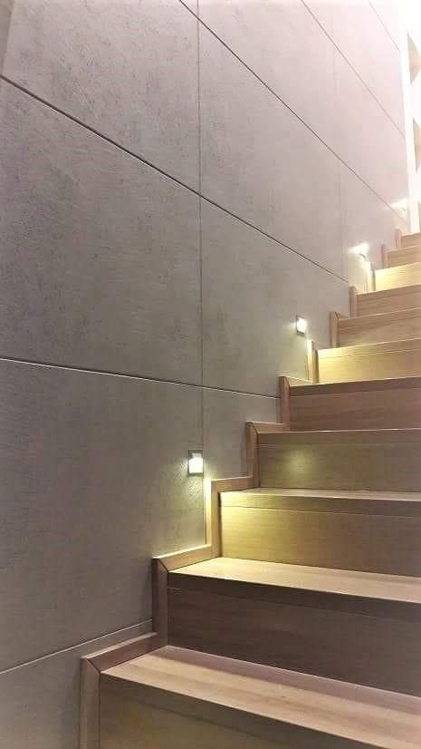 Nowoczesne schody drewniane z podświetleniem LED wbudowanym w ścianę z szarych paneli imitujących beton.