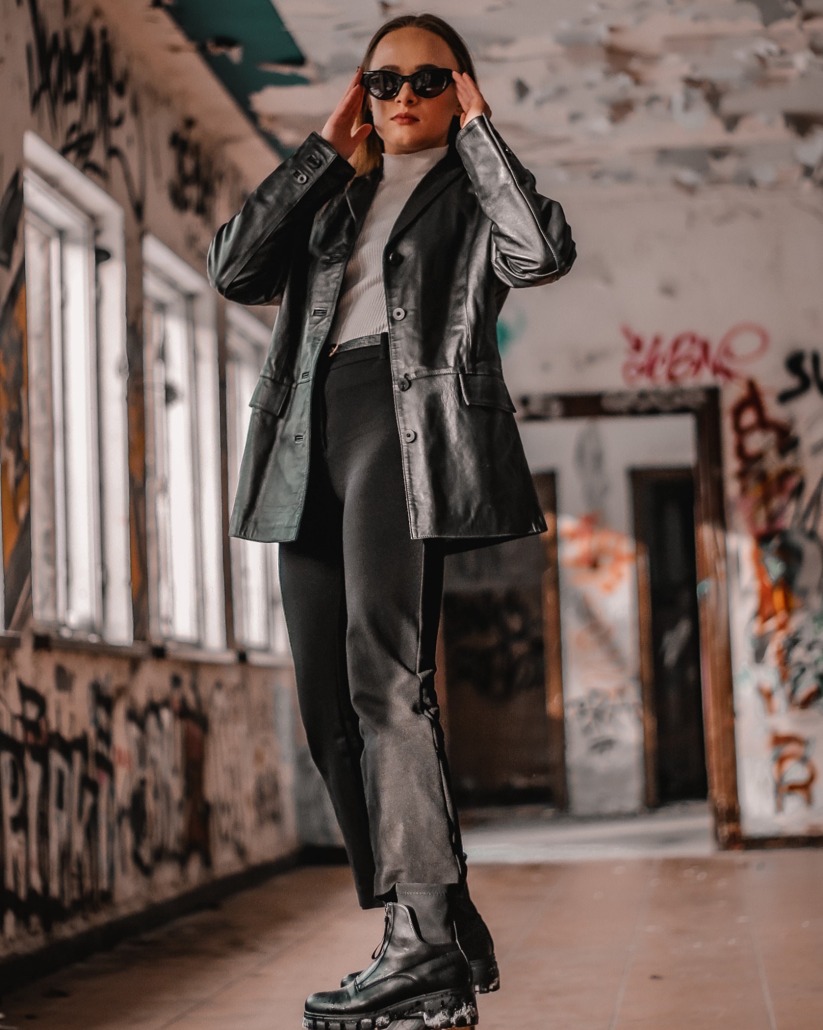 Kobieta w czarnej skórzanej marynarce i okularach przeciwsłonecznych pozuje w opuszczonym budynku z graffiti na ścianach.