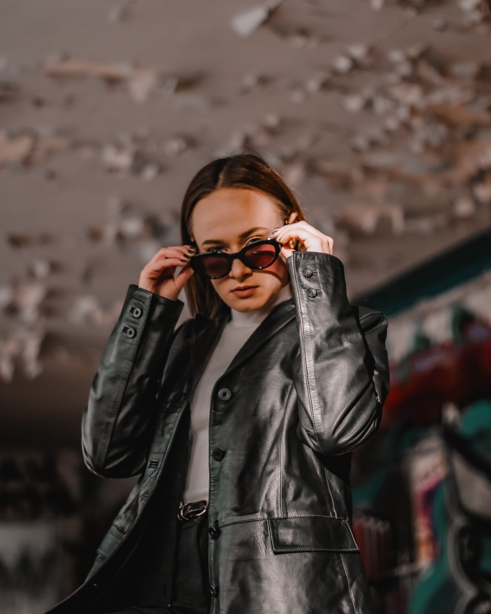 Kobieta w czarnej skórzanej marynarce i okularach przeciwsłonecznych, pozująca na tle ściany z odpadającą farbą i graffiti.