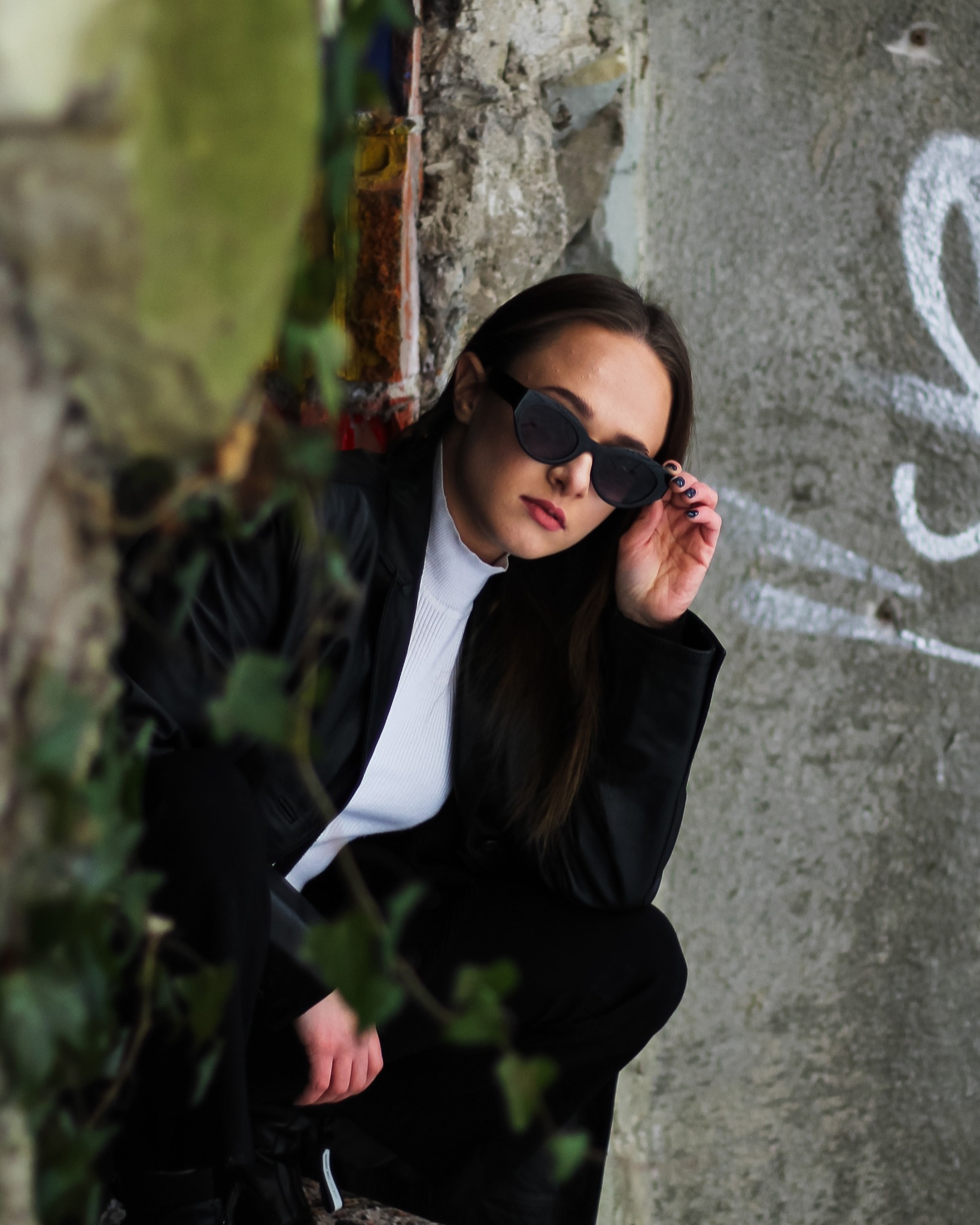 Kobieta w czarnym garniturze i okularach przeciwsłonecznych, kuca przy betonowej ścianie z graffiti i bluszczem, pozując do zdjęcia
