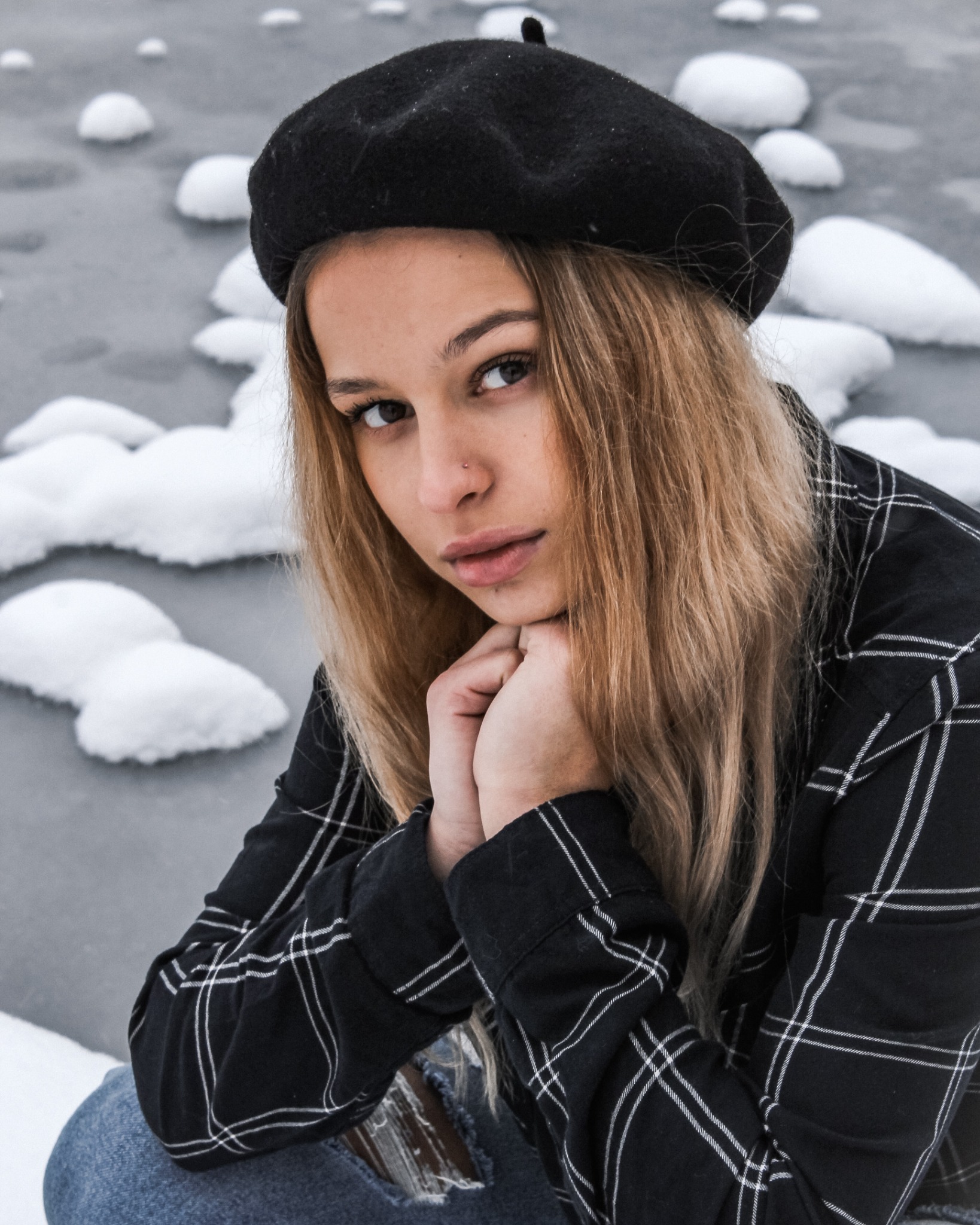 Portret młodej kobiety w czarnym berecie i koszuli w kratę, opierającej brodę na dłoniach, na tle zimowego krajobrazu z plamami śniegu.