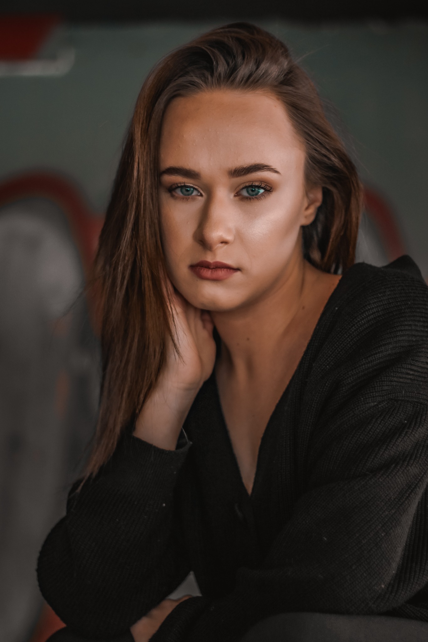 Portret młodej kobiety z długimi brązowymi włosami i jasnymi oczami, opierającej podbródek na dłoni, ubrana w czarny sweter na tle szarej ściany z abstrakcyjnymi czerwonymi wzorami.